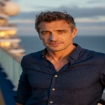 **middle-aged man in his late 40s, fit build, slightly weathered yet attractive face, gray-green eyes with calm intensity, short tousled dark brown hair with subtle silver streaks, light tan skin, dressed in a fitted blue button-up shirt with sleeves rolled, open collar, relaxed but confident posture, standing on the deck of a cruise ship at sunset, soft warm lighting, sea breeze moving his hair, cinematic realism, shallow depth of field, 85mm portrait photography style --ar 2:3 --v 7.0** - Image #3 <@876267919003238460>