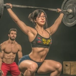 **a photo of an asian woman in her thirties doing an overhead squat wearing a black shirt with butterfly dream motif and blue shorts at the gym. She is wearing glasses, and there are some boxes on the ground, weight plates around her, and another person next to her observing how she performs the exercise. the setting is a dark room with a concrete floor and a barbell in the background. She looks focused while lifting heavy weights overhead. the image captures a moment during a crossfit training session. --p pu1nj44** - Image #3 <@538878031226470440>