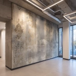 **A photo of an intricate, hand-painted wallpaper with faded gold and silver designs on the wall in an office building. The texture is rough concrete with visible sanded edges. There's an LED light bar above it that casts a soft glow over its surface. A windowless corner wall is on one side. In the background, there's another wallpaper design that looks similar but less detailed. It has more colors and patterns. The room also features white square lighting fixtures on the walls, creating subtle shadows on the ceiling. --ar 32:23** - Image #2 <@1313000520759377972>