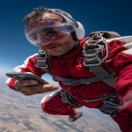 **The photo should be hyperrealistic but with distorted proportions. The framing should distort the proportions, prioritizing the head and shrinking the legs and feet. A 40-year-old man is skydiving, having fun, and wearing clear protective glasses. He's holding a smartphone in his right hand and his gaze is directed at the screen. Focus on the arm and the phone in the foreground. Use of a wide-angle lens. Natural light, surreal photo, crazy proportions. --ar 9:16** - Image #4 <@1310560084346146826>