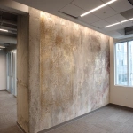 **A photo of an intricate, hand-painted wallpaper with faded gold and silver designs on the wall in an office building. The texture is rough concrete with visible sanded edges. There's an LED light bar above it that casts a soft glow over its surface. A windowless corner wall is on one side. In the background, there's another wallpaper design that looks similar but less detailed. It has more colors and patterns. The room also features white square lighting fixtures on the walls, creating subtle shadows on the ceiling. --ar 32:23** - Image #1 <@1313000520759377972>