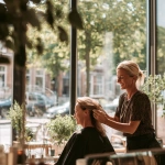 **a smiling female hairdresser in her early forties is standing arranging the hair of a client in a beauty salon facing the large mirrors, beside the windows that give on a lovely street full of green treas. It's a beautiful day and the warm sunlight enters the beauty salon through the windows, 35 mm, realistic, --s 50 --v 7.0 --raw** - Image #2 <@1088739679458824253>