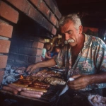 **grumpy Portuguese mustachio'd old man grilling delicious barbecue assorted meats, sausages and chicken over hot coals in a brick barbecue oven, wiping the sweat off his brow with tiny towel, with tanabata-inspired print shirt, photo taken with provia** - Image #2 <@538878031226470440>