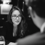 **Black and white candid DSLR photo of a dinner scene. A young woman about 27 sits at the table, wearing glasses and an elegant secretary outfit. She has long flowing hair and a natural realistic face, with small imperfections and a subtle hidden smile as she looks at the man across from her. Her posture is relaxed and graceful, expressing warmth and presence. The man is blurred in the foreground, only partly visible. The image is simple, authentic, like an unedited real photograph.** - Image #3 <@1223901074742837300>