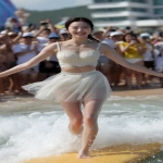 **a joyful Korean university girl with pale glowing skin, camera-facing with a wide smile, standing tall on a surfboard in motion, wearing an ivory sweetheart neckline crop top with lace trims and a translucent soft grey ballet mini skirt, toes visible with soft wet sand, beach crowd cheering behind --v 7.0 --ar 9:16 --q 2 --raw** - Image #4 <@1192072662005059597>