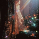 **1970s film grainy photograph. A twinkling waterfall. Big statues of freestanding walls, covered with black tiny rainbow colored holographic rhinestones glinting in the sun. Around the statue is a fashion model, some curvy plus-size, some of different ethnicities. She wears an opulent gothic futuristic pink and blue, satin lace and fish-net outfit, adorned with roses. Eerie sci-fi aesthetic. Bright colorful light. sparkly airy misty twinkles fall from the sky. Orange, peach, pink, blue, lime green, purple --v 7.0** - Image #1 <@840730232632639528>