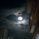 **A photograph of the moon in a black sky, taken from an apartment balcony with a striped, patterned awning and flat white walls, seen through a window of another dark building. The photograph is taken at night. There are some clouds in front of the moon, covering up most of the full moon. It looks like real smartphone footage. --ar 3:4 --raw** - Image #2 <@699706172105556009>