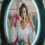 **A medium shot of a stylish Western female fashion influencer livestreaming, actively holding a white lace blouse against her body to demonstrate the fit, her hands are visible gripping the hanger and touching the fabric details to showcase the texture. She has an exaggerated shocked and excited expression with her mouth open. In the foreground, a blurred silhouette of a ring light and smartphone recording the scene. Background features a rack of colorful clothes. High energy, commercial livestream photography, sharp focus on the influencer and the clothing, with high detail ,photograph --ar 16:9** - Image #2 <@950232969275125781>