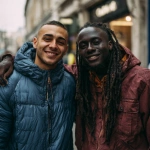 **a photo of two handsome young men smiling and posing for the camera. one is white with short brown hair wearing a blue puffer jacket, the other is a black man with long dreadlocks on top and a fade haircut, wearing a dark red-orange windbreaker. the background is an urban street setting with buildings and signs, and they both have their arms around each other's shoulders, in the style of annie leibovitz. --ar 127:128 --v 7.0 --p 7t8d38w** - Image #4 <@995751013732581408>