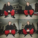 **A young handsome tan skinned white man with dirty blonde wavy short hair dressed in a Y2K Cholo gangster style, wearing a black long sleeve Dickies work shirt and extremely baggy, extra-long, 3XL plain long red and black Nike basketball shorts. His shorts are very long and go down to his ankles and feet. He is wearing Nike Cortez shoes in his house by a Christmas tree** - Variations (Strong) by <@1061360796438900746> (fast)