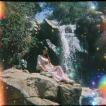 **1970s film grainy photograph. A twinkling waterfall. Big statues of freestanding walls, covered with black tiny rainbow colored holographic rhinestones glinting in the sun. Around the statue is a fashion model, some curvy plus-size, some of different ethnicities. She wears an opulent gothic futuristic pink and blue, satin lace and fish-net outfit, adorned with roses. Eerie sci-fi aesthetic. Bright colorful light. sparkly airy misty twinkles fall from the sky. Orange, peach, pink, blue, lime green, purple --v 7.0** - Image #1 <@840730232632639528>