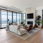 **ultra-realistic photograph of a coastal modern open floor plan living room in Manhattan Beach, California, warm oak wide plank hardwood floors, large black-framed windows with panoramic ocean and beach view, Restoration Hardware modern-inspired neutral palette, plush L-shaped sectional rotated so one side faces the ocean and the other faces a sleek TV above a limestone fireplace, layered grass cloth and Persian area rug, stone coffee table, leather lounge chair, wood rectangle couch table with lamp and fresh flowers and books on top, textured throw pillows, soft ambient natural daylight, sculptural chandelier, layered organic textures, styled for a luxury interior design Instagram post, straight-on wide-angle lens, full view of space including ocean view and TV wall, center fireplace, remove table next to chair, photorealistic details, depth, natural shadows --v 6.0 --raw --ar 13:10** - Image #4 <@1339375378858381312>