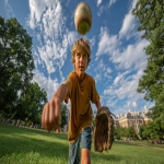**Image from slightly above eye level: a 10-year-old boy wearing blue shorts and an ochre t-shirt stands on a lush green park lawn, mid-action as he throws a baseball directly toward the viewer. A baseball glove is on his hand, and the ball floats mid-air above him. He is looking directly at the camera. Natural sunlight with soft shadows illuminates the scene. The background features a clear blue summer sky, distant trees, and buildings along the horizon. Full-body view, professional color grading, clean sharp focus, no grain, cinematic quality. Shot with a Sony Alpha A7 III and Zeiss ultra-macro lens, f/2.8 aperture, high resolution:: --aspect 16:9 --s 250** - Image #1 <@1220830039520841890>