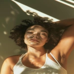 **portrait of a young mixed-race woman with curly hair, slightly dewy skin, soft freckles, natural lips, relaxed facial expression, ambient natural light casting shadows across her face, golden hour glow, shot from below at a close angle, wearing a minimal tank top, realistic skin texture, subtle pores, no makeup look, candid and intimate atmosphere, real human face proportions, 35mm lens look, high-detail photography --v 6.0 --ar 2:3 --no airbrushed skin, no artificial eyes, no smooth plastic texture --raw** - Image #2 <@693446385436983346>