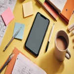 **a photograph of an iphone on a clean work desk in the office with a binder full of papers on the desk, a notepad and pen, stationery. two post-its on the wall. a half-drank coffee mug. the phone should take up about half the visual space. the photography style is colourful and editorial, colour palette should be warm and inviting. <https://s.mj.run/dF8N_y-B2KE> --ar 16:9** - Image #4 <@676493813367177236>