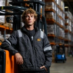 **A male forklift specialist in bright workwear and a helmet, with curly hair and an expressive gaze, standing next to a forklift, leaning on it. In the background, there is a warehouse filled with products neatly arranged on shelves. The scene is softly lit, highlighting the details of his outfit, the forklift, and the products, creating an atmosphere of professionalism, confidence, and a work-focused vibe. --p ud5ebxz** - Image #1 <@1073058804779978844>