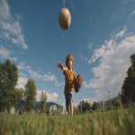 **Image from slightly above eye level: a 10-year-old boy wearing blue shorts and an ochre t-shirt stands on a lush green park lawn, mid-action as he throws a baseball directly toward the viewer. A baseball glove is on his hand, and the ball floats mid-air above him. He is looking directly at the camera. Natural sunlight with soft shadows illuminates the scene. The background features a clear blue summer sky, distant trees, and buildings along the horizon. Full-body view, professional color grading, clean sharp focus, no grain, cinematic quality. Shot with a Sony Alpha A7 III and Zeiss ultra-macro lens, f/2.8 aperture, high resolution:: --aspect 16:9 --s 250** - Image #3 <@1220830039520841890>