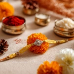**A close-up photograph of a festive Raksha Bandhan setup on a soft beige background. A delicate yellow thread rakhi with silver bead accents is placed in the center, surrounded by a vibrant orange marigold flower, a small white jasmine flower, metallic silver containers filled with red kumkum and rice, and a pinecone for added texture. The lighting is warm and soft, giving a cozy and elegant festive feel, with a shallow depth of field that keeps the focus on the rakhi.** - Image #2 <@1331627685406965792>