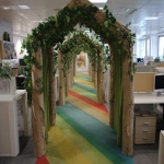 **The tunnel structure is made of thin support poles from a garden arch measuring 1.5 meters in height, 1.2 meters in width, and 5 meters in depth. Inside the entrance of the cardboard arch, the exterior is decorated with a forest interior, and a path made of thin rainbow-colored vinyl tape is depicted on the floor.a cotton cloud canopy, and an interior resembling library shelves mapped onto cardboard at the end of the forest. A children's attraction interacting with a server station is installed by covering office desks and displays with fabric, placing them in the pathways between desks, removing desk chairs to create pathways, and masking the lower parts of desks with green curtains. Cardboard guardrails are installed up to waist height, creating a bright and cheerful atmosphere. --ar 4:3** - Image #1 <@580349603761750038>